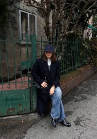 Γυναίκα που φοράει μαύρο παλτό, μπλε τζιν, μαύρο καπέλο και παπούτσια κάθεται σε τοιχίο από τούβλα δίπλα σε πράσινο μεταλλικό φράκτη σε δρόμο.