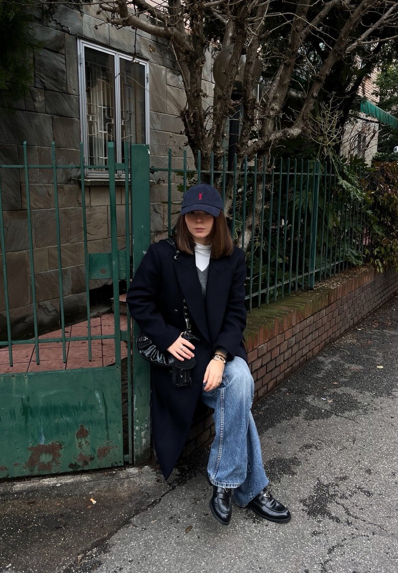 Kvinne iført svart kåpe, blå jeans, svart caps og sko sitter på en mursteinshylle ved siden av et grønt metallgjerde på en gate.