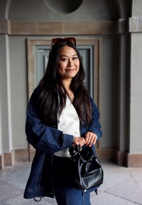 Denim jacket with button closure, layered over a white top. Black leather handbag with gathered accents, held in hands. Dark hair, neutral background.