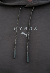 Le sweat à capuche gris comporte un cordon de serrage avec des extrémités texturées, affichant "HYROX" et un logo Puma dans une couleur plus claire contrastante.