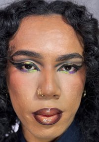 Zbliżenie osoby z kręconymi czarnymi włosami, odważnym, wielokolorowym makijażem oczu, złotymi kolczykami w nosie i błyszczącą metaliczną miedzianą szminką.