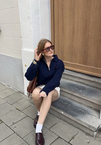 Kvinna i solglasögon, marinblå zip-up-tröja, vita shorts och brun väska som sitter på utomhustrappan vid en träddörr.