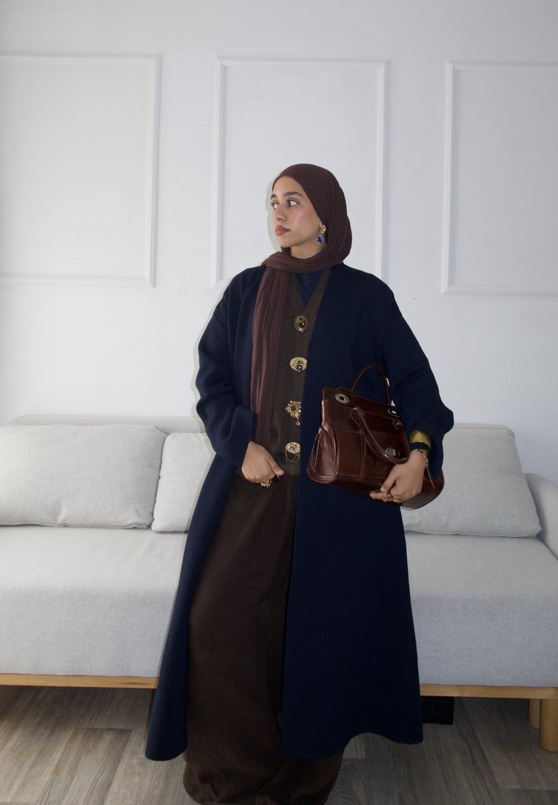 Lungo cappotto blu navy, abito marrone con dettagli a bottoni, sciarpa marrone, in possesso di una borsa marrone. Design semplice su uno sfondo chiaro con un divano.