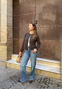 Ciemnobrązowy krótki kardigan na białym topie, do tego jasnoniebieskie dzwony. Trzyma beżową torbę, na tle teksturowanego brązowego tła.