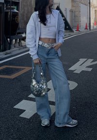 Ανοιχτό γκρι φούτερ με φερμουάρ, λευκή κοντή μπλούζα, ανοιχτό μπλε παντελόνι με φαρδιά πόδια, ασημένια ζώνη με στρας, λαμπερή τσάντα, ασημένια sneakers, αστικός φόντο.