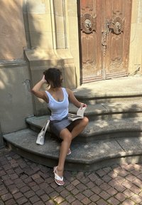 Žena v bielom tielku a šedých sukni sedí na kamenných schodoch, číta knihu, vedľa seba má bielu kabelku a za ňou je zdobené drevené dvere.