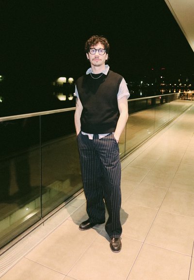 Joven con gafas, pelo rizado, lleva un chaleco negro, pantalones de rayas y mocasines, está de pie en un balcón por la noche con las luces de la ciudad reflejadas.