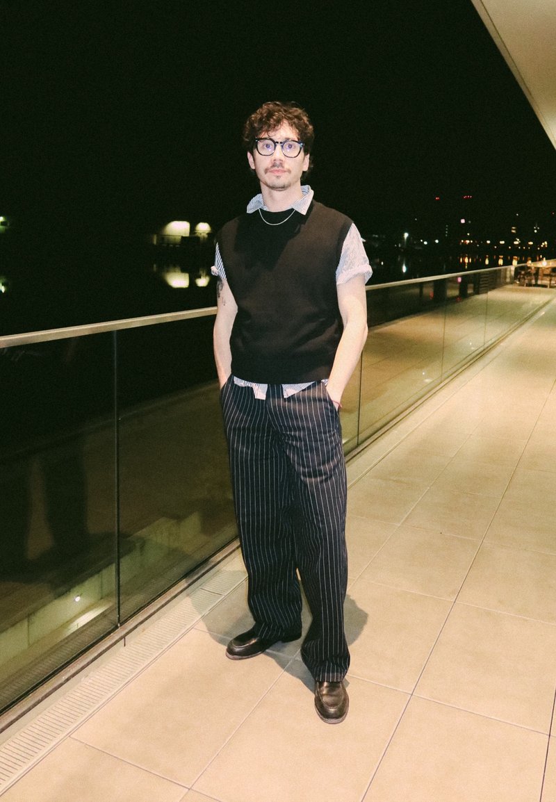 Νέος άνδρας με γυαλιά, μαλλιά σγουρά, φοράει μαύρο γιλέκο, ριγέ παντελόνια και μοκασίνια, στέκεται σε μπαλκόνι τη νύχτα με αντηχήσεις φώτων της πόλης.