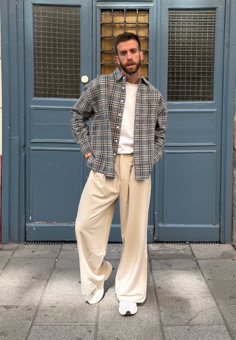 Camicia flanella a quadri grigia e nera sopra una camicia bianca, abbinata a pantaloni beige a gamba larga e sneaker bianche davanti a uno sfondo di porta blu.