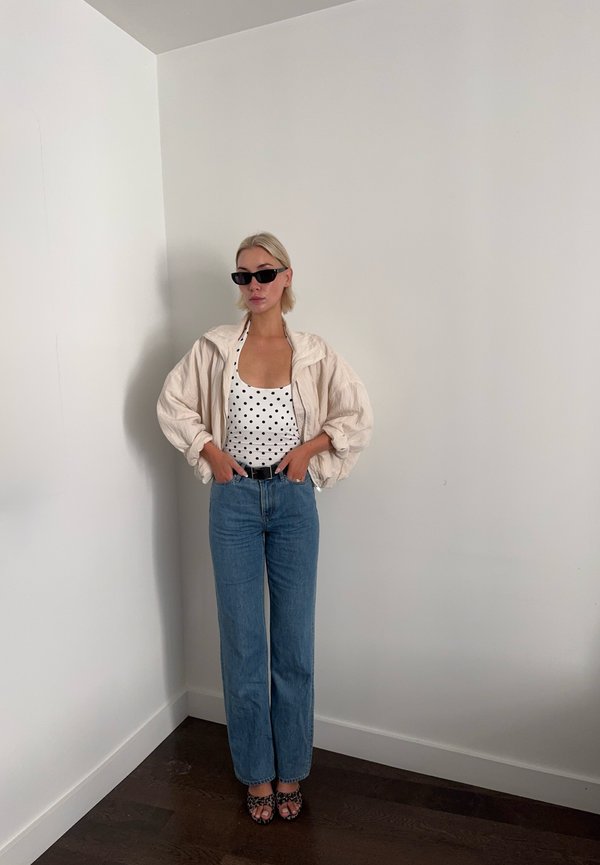 Femme portant des lunettes de soleil noires, un haut blanc à pois, une veste beige clair, un jean bleu et des chaussures imprimé léopard, debout contre un mur blanc.
