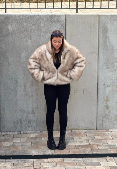 Chaqueta corta de piel sintética beige con cremallera frontal, llevada sobre una camiseta negra y leggings negros. Combinada con botas de combate negras con cordones.