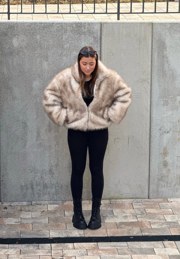 Veste courte en fausse fourrure beige avec fermeture éclair à l'avant, portée sur un haut noir et des leggings noirs. Associée à des bottes de combat noires à lacets.
