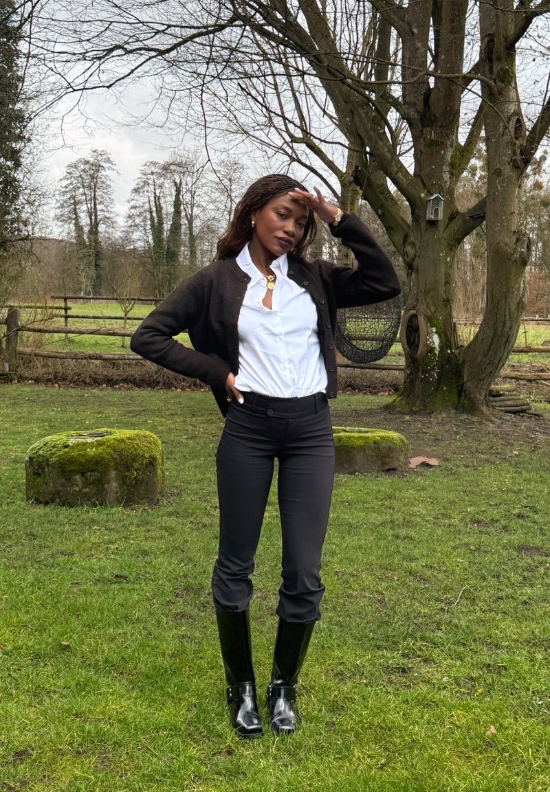 Femme en cardigan noir, chemise blanche, pantalon noir et bottes hautes noires, debout sur l'herbe près de grands arbres et de pierres recouvertes de mousse dans un cadre rural.