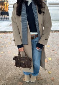 Beige trench kabát, sötétkék kardigán, csíkos sál, kék farmernadrág és világosbarna slip-on cipő. Sötétbarna bőr kézitáska cipzár részlettel.