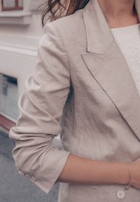 Lichtbeige linnen blazer met een getailleerde pasvorm, notch-revers en drie knopen op de manchetten. Gestructureerde stof is zichtbaar.