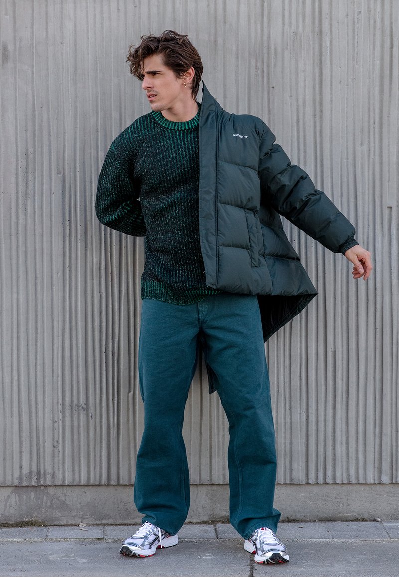 Tamsiai žalia pūkinė striukė ant tekstūruoto megztinio, derinama su žaliais kelnėmis ir baltomis sportinėmis avalynėmis. Pilka fono spalva su vertikaliomis linijomis.