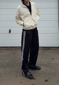 Beige lightweight zip-up jacket with a textured pattern, black polo shirt underneath, black track pants with white stripes, and black slip-on shoes.