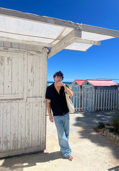 Camisa negra de manga corta, jeans azul claro y una bolsa tote beis. Cerca de una estructura de madera desgastada y cabañas de playa coloridas bajo un cielo despejado.