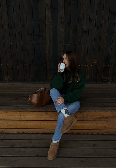 Botas de ante marrón hasta el tobillo, jeans azules, sudadera verde, sosteniendo una taza blanca. Bolsa de cuero marrón sobre escalones de madera contra un fondo de madera oscura.
