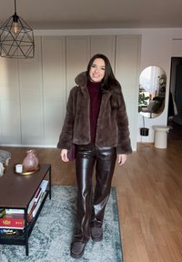 Femme souriante à l'intérieur portant une veste en fausse fourrure marron, un col roulé bordeaux, un pantalon en cuir foncé et des chaussures assorties marron, debout sur un tapis.