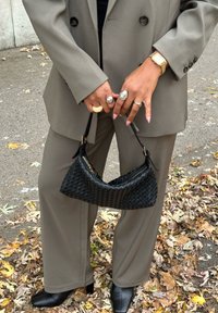 Bolso tejido negro sostenido por las manos, combinado con un blazer oversize color cuero y pantalones a juego. Se ven dedos con anillos y botas negras de tacón.