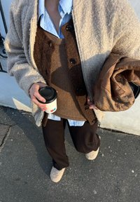 Μπεζ φλις ζακέτα πάνω από ένα καφέ πλεκτό γιλέκο, στρωμένο πάνω από ένα ανοιχτό μπλέ πουκάμισο. Σκούρες ριγέ παντελόνες και καφέ φλις Παντόφλες. Κρατώντας μια κούπα καφέ.