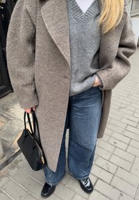 Cappotto di lana grigio, maglione grigio a scollo a V, jeans a gamba larga blu, borsa nera e scarpe nere lucide su pavimentazione testurizzata.
