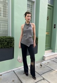 Haut sans manches gris avec un col drapé, associé à un pantalon noir évasé. Tient un sac à main en filet. Mur vert et plantes en pot en arrière-plan.
