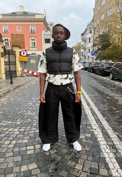Chaleco de plumas negro sobre una camisa blanca con estampado de aves, pantalones negros holgados y zapatos blancos. De pie en una calle empedrada con edificios al fondo.