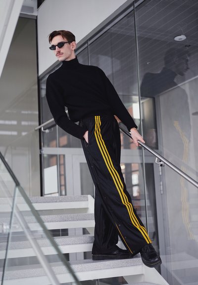 Jersey de cuello alto negro combinado con pantalones negros brillantes con rayas laterales amarillas, de pie en unas escaleras con barandilla de cristal, usando gafas de sol.