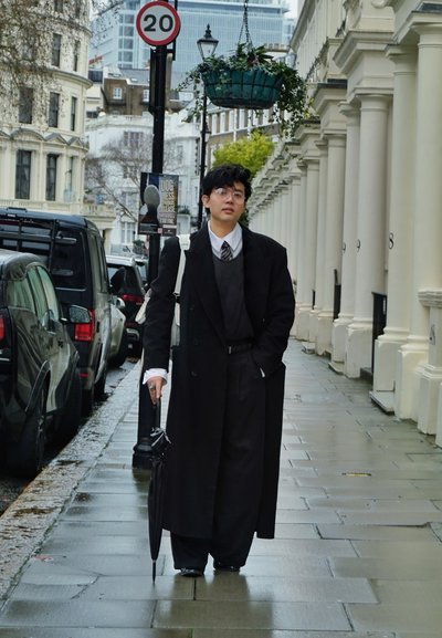 Persona con abrigo largo negro y gafas, sosteniendo un paraguas, caminando por una acera mojada de la ciudad cerca de coches estacionados y edificios con columnas.