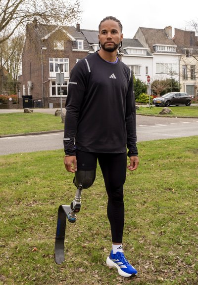 Atleta con camiseta de manga larga negra y leggins de pie sobre la hierba. Notable pierna prostética de fibra de carbono y zapatillas deportivas azules.