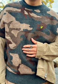 Grau-brauner Camouflage-Pullover mit rundem Halsausschnitt, aus strukturiertem Material gefertigt, mit einer gelben Logo-Stickerei. Hände zeigen silberne Ringe.
