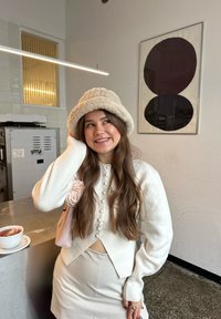 Jeune femme en tricot blanc et chapeau seau en peluche souriant debout près d'un comptoir avec une tasse de thé dans un café moderne.
