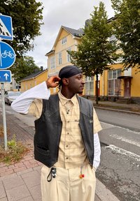 Svart läderväst över en rutig beige skjorta, kombinerat med kremfärgade byxor. Accessoarer inkluderar en kedja och färgglada nyckelringar. Urban bakgrund.