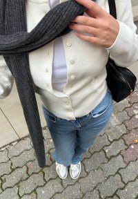 Bela kardigan s gumbi, položen preko bele majice, v kombinaciji z modrimi džinsom in srebrnimi supergami. Temno sivi rebrasti šal dopolnjuje videz.