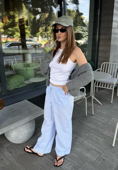 Top sin mangas blanco, cárdigan gris, pantalones anchos de rayas en azul claro, chanclas negras, gorra verde y gafas de sol; sosteniendo una bebida verde.