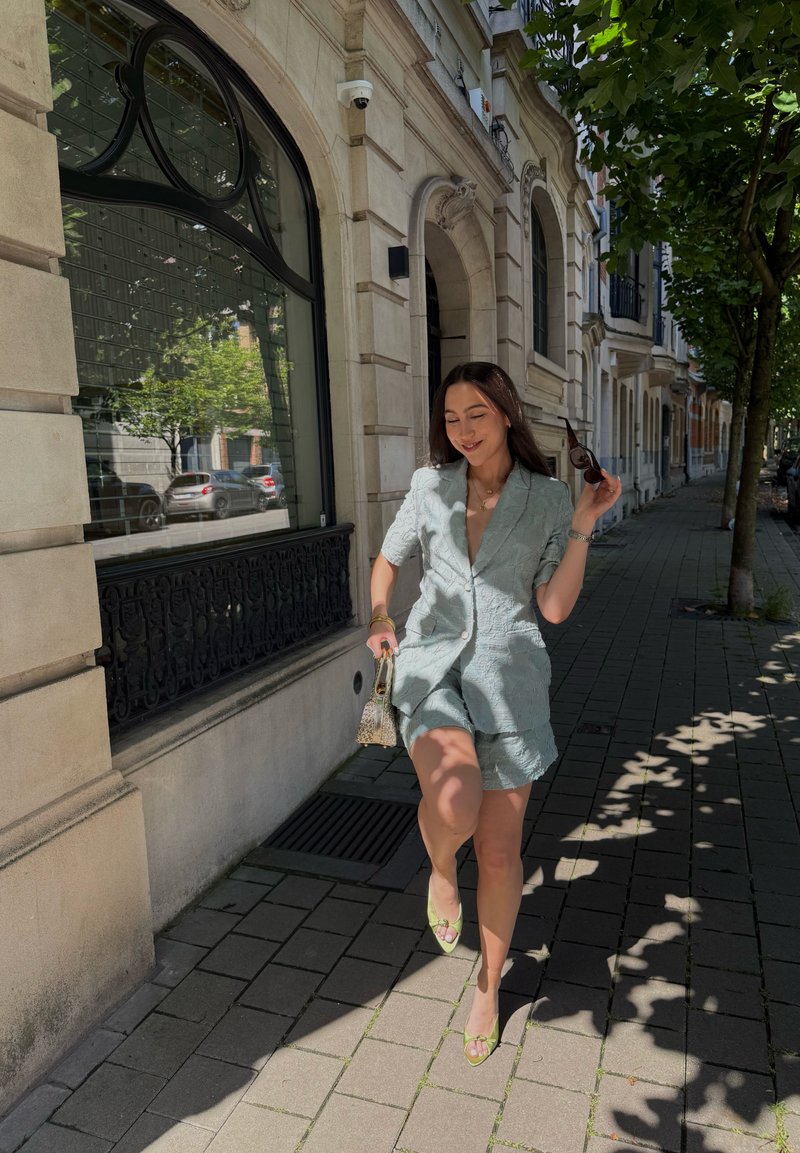 Femme portant un blazer bleu clair et un short, tenant des lunettes de soleil et un petit sac, marchant sur un trottoir ensoleillé avec des arbres et des bâtiments à proximité.