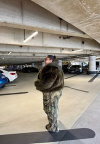 Veste en fausse fourrure vert foncé, oversize, avec capuche. Pantalon camouflage aux nuances de vert et de brun. Fond de parking en béton.