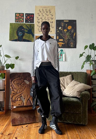 Pantalones cargo elásticos negros, camiseta de manga larga blanca con tres franjas, chaqueta negra y zapatillas negras con franjas blancas, de pie cerca de un sofá verde.