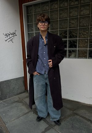 Persona con cabello castaño corto y gafas grandes lleva un abrigo largo oscuro, camisa azul, vaqueros sueltos y zapatos negros, de pie junto a una pared de bloques de cristal.