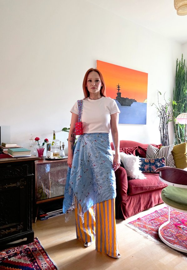 Mujer de pie en una sala de estar colorida, vistiendo una camisa blanca, falda cruzada azul con flores y pantalones amarillos a rayas con un bolso rojo.