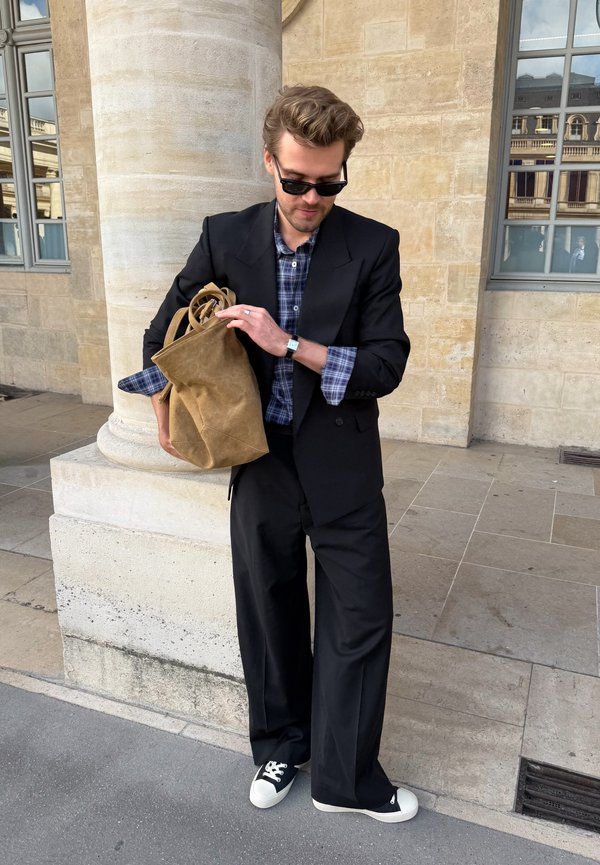Hombre con traje negro y gafas de sol sostiene una gran bolsa marrón, de pie cerca de una columna de piedra y una ventana de un edificio beige.
