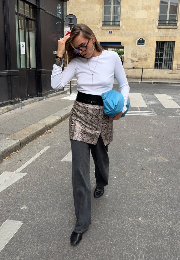 Une femme portant des lunettes traverse une rue en ville, un sac bleu à la main, et écarte ses cheveux du visage.