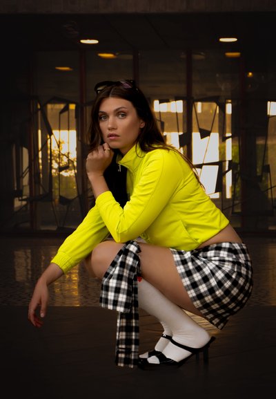Chaqueta corta neón amarilla, falda de cuadros en blanco y negro, medias blancas por la rodilla, tacones negros y cabello largo castaño. Entorno urbano moderno.