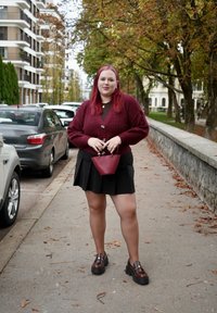 Bordový pletený sveter cez hnedý top, čiernu sukňu a hnedé platformové topánky; drží malú bordovú kabelku. Jeseň v pozadí.