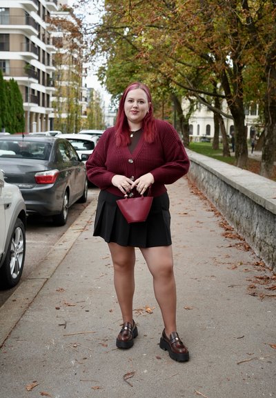 Cárdigan de punto burdeos sobre una blusa marrón, falda negra y zapatos de plataforma marrones; sosteniendo un pequeño bolso burdeos. Hojas de otoño en el fondo.