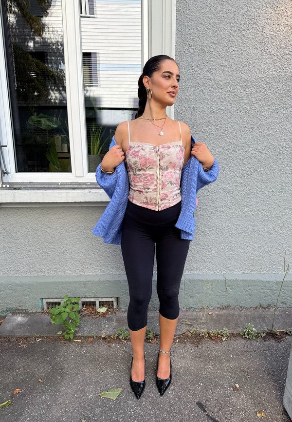 Donna con capelli intrecciati che indossa un top corsetto floreale, un cardigan blu, leggings neri corti e tacchi neri, in piedi su un marciapiede accanto a un muro grigio.