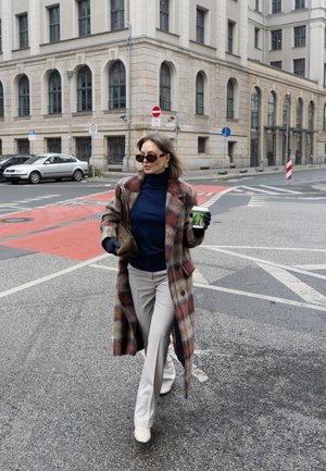 Mujer con abrigo de cuadros, gafas de sol, pantalones beige y guantes sostiene una taza de café mientras cruza una calle de la ciudad cerca de edificios históricos.