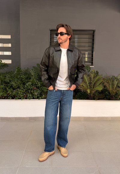 Hombre con chaqueta de cuero negra, camisa blanca, vaqueros azules y zapatos beige, de pie con las manos en los bolsillos cerca de plantas y una ventana.